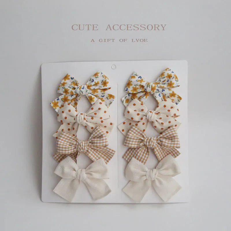 Set of eight children's cloth bow hair clips in various colours including beige polka dot, pink, blue floral, and wine red plaid patterns.