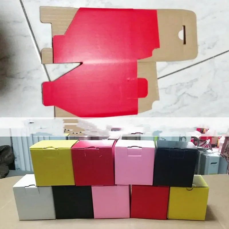 Square corrugated cardboard box in black, shown next to white and grey boxes, with a ruler indicating 10cm size.