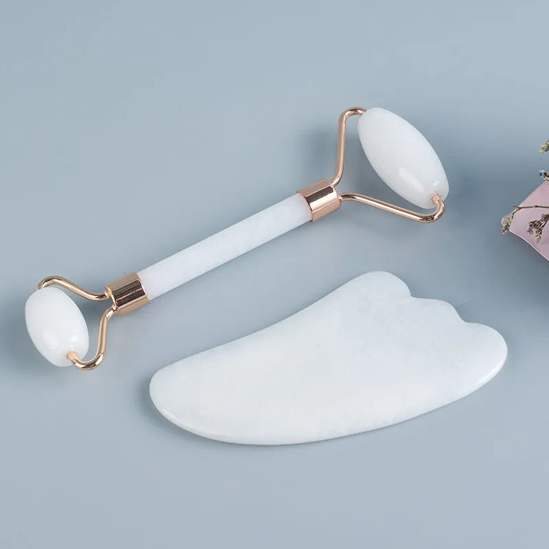White jade facial roller and gua sha scraper board set on a plain surface.