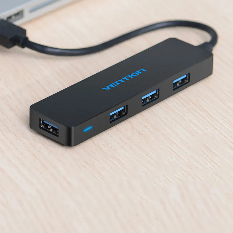 Black USB 3.0 four-port hub with a short cable and nickel-plated connectors on a desk.