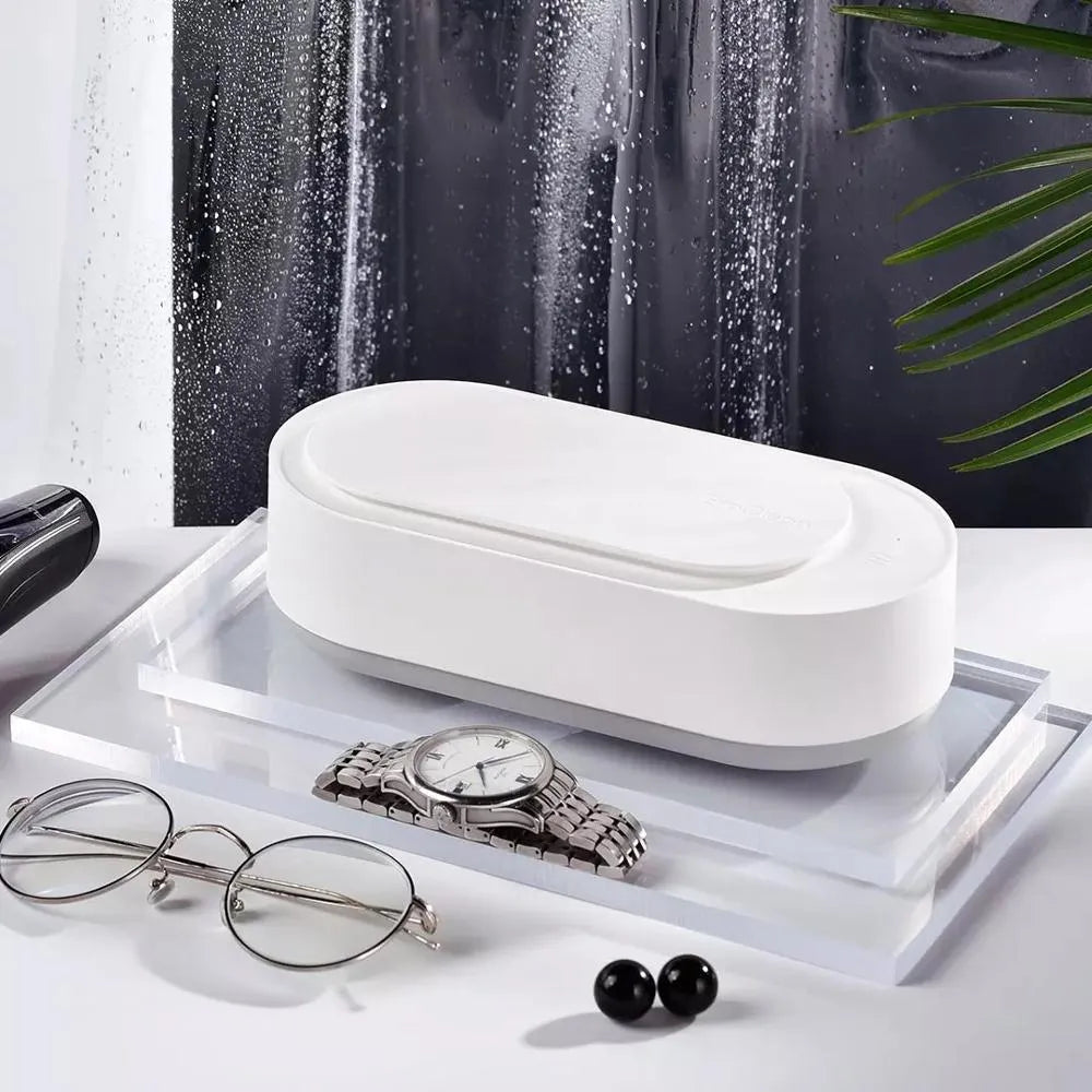 White portable ultrasonic cleaner with touch button next to jewellery and glasses on a table.