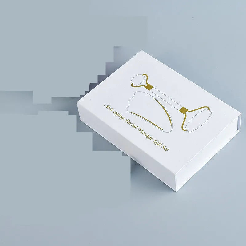 White jade facial roller and gua sha scraper board set on a plain surface.