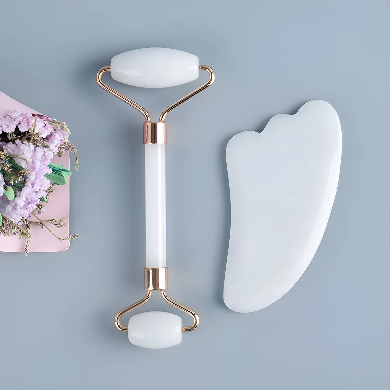 White jade facial roller and gua sha scraper board set on a plain surface.