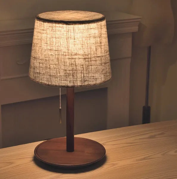 Walnut coloured solid wood bedside lamp with a white cloth shade on a wooden table.