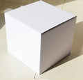 Square corrugated cardboard box in black, shown next to white and grey boxes, with a ruler indicating 10cm size.