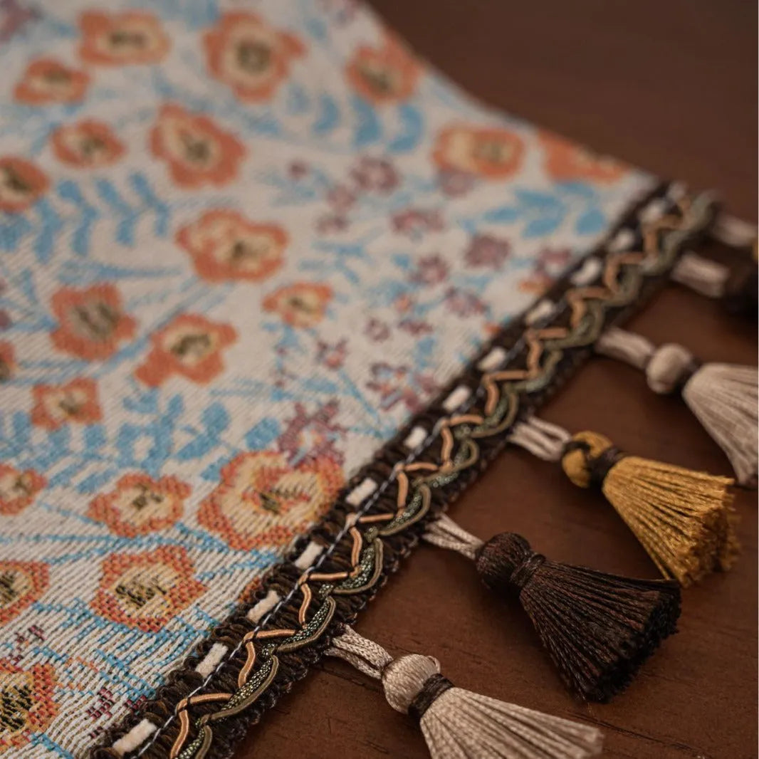 Beige jacquard table runner with floral pattern and long tassels laid on a wooden table.