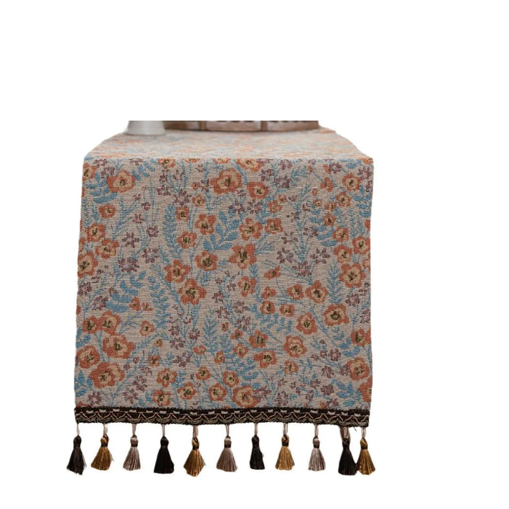 Beige jacquard table runner with floral pattern and long tassels laid on a wooden table.