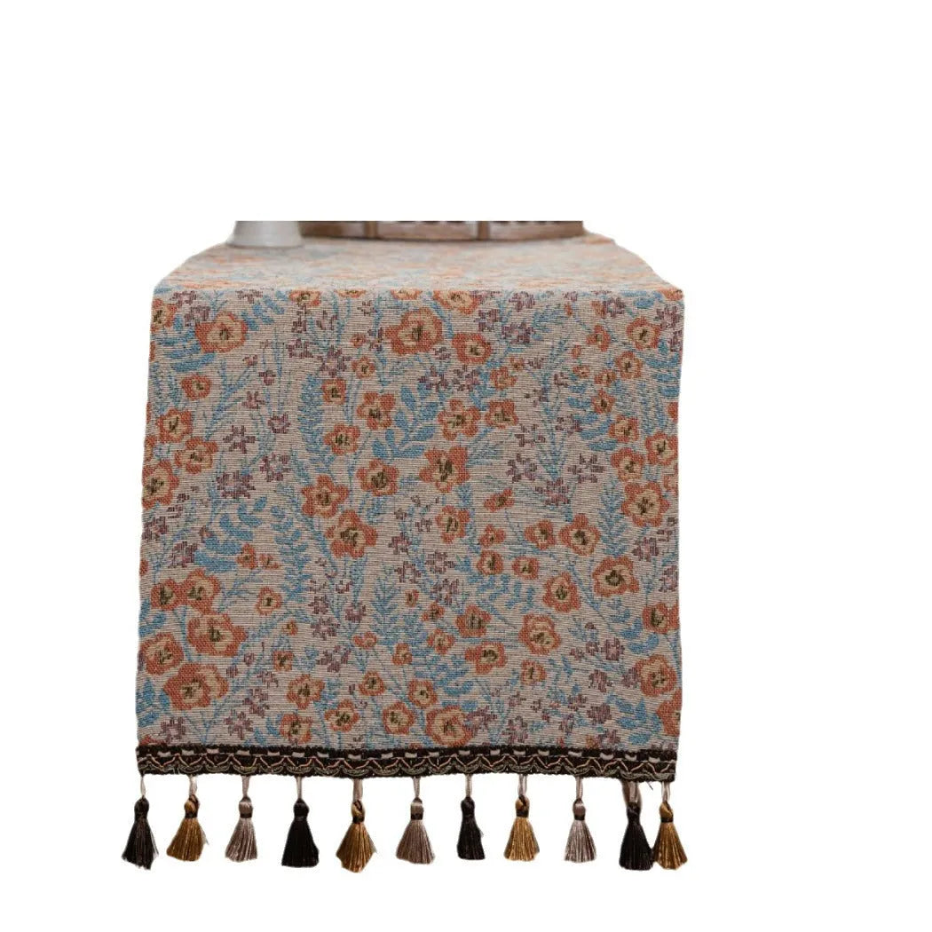 Beige jacquard table runner with floral pattern and long tassels laid on a wooden table.