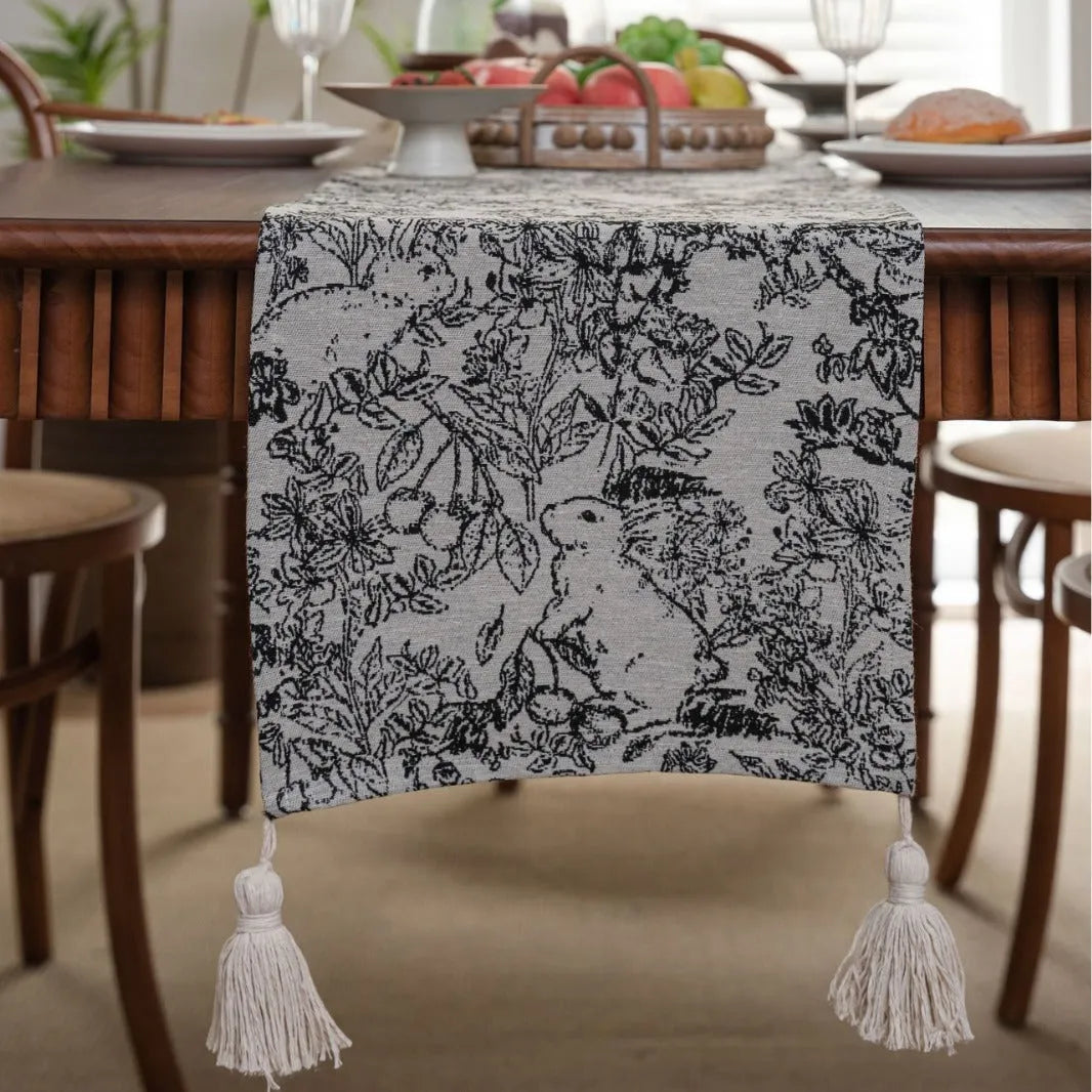 Beige jacquard table runner with floral pattern and long tassels laid on a wooden table.