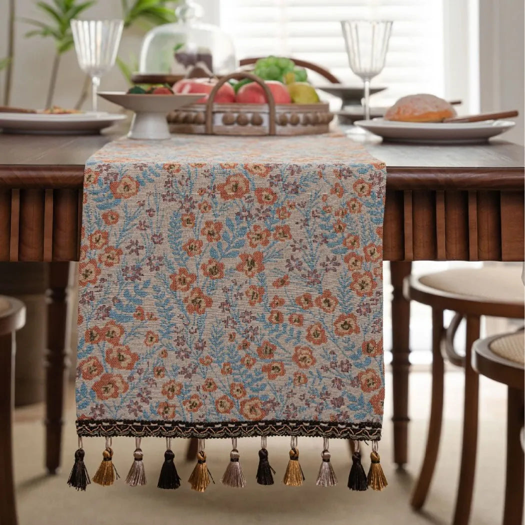 Beige jacquard table runner with floral pattern and long tassels laid on a wooden table.