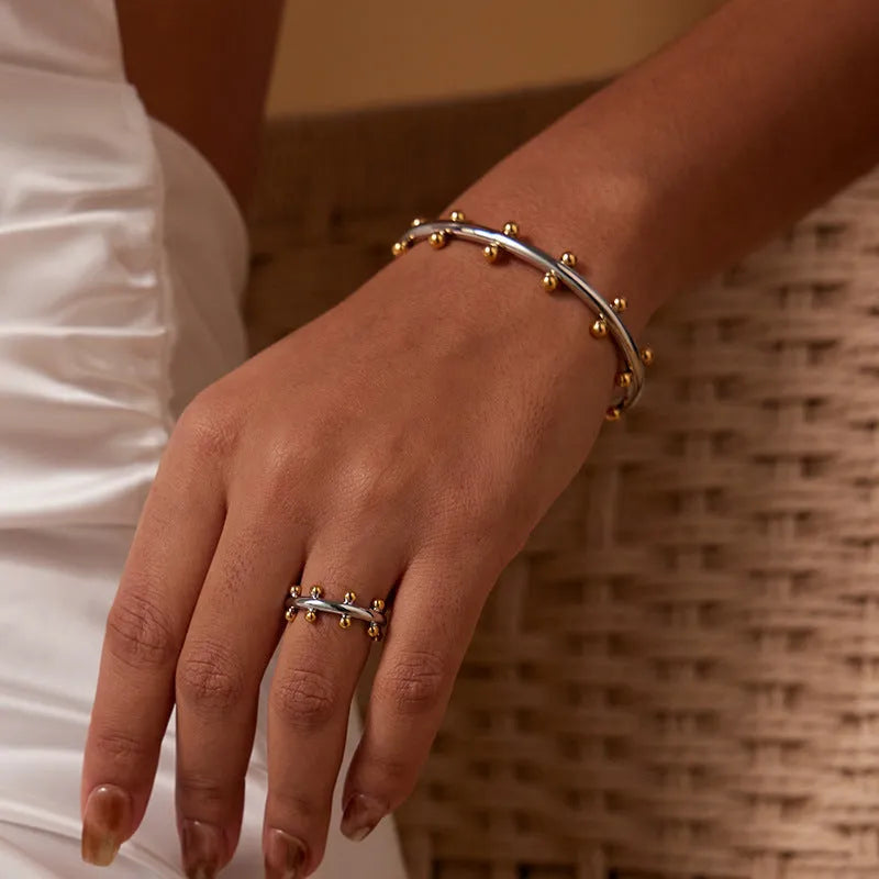 Close-up of textured stainless steel C-shaped bean bead charm in gold finish, shown next to an alloy bracelet.