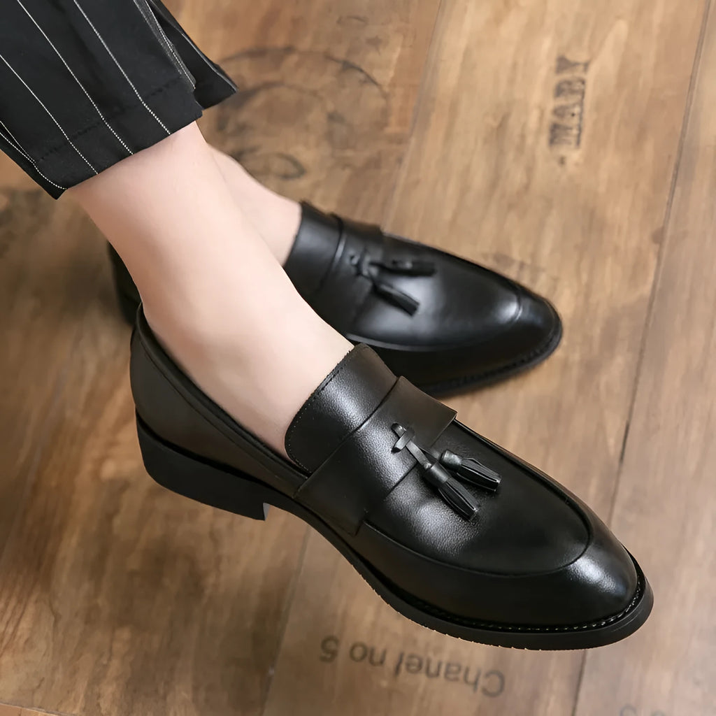 Men's black tassel slip-on loafers with a pointed toe and rubber sole on a white background.