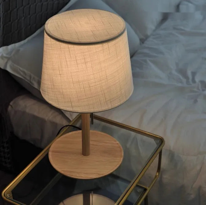 Walnut coloured solid wood bedside lamp with a white cloth shade on a wooden table.