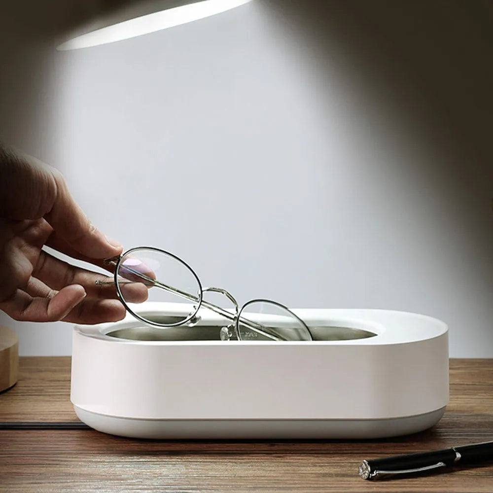 White portable ultrasonic cleaner with touch button next to jewellery and glasses on a table.